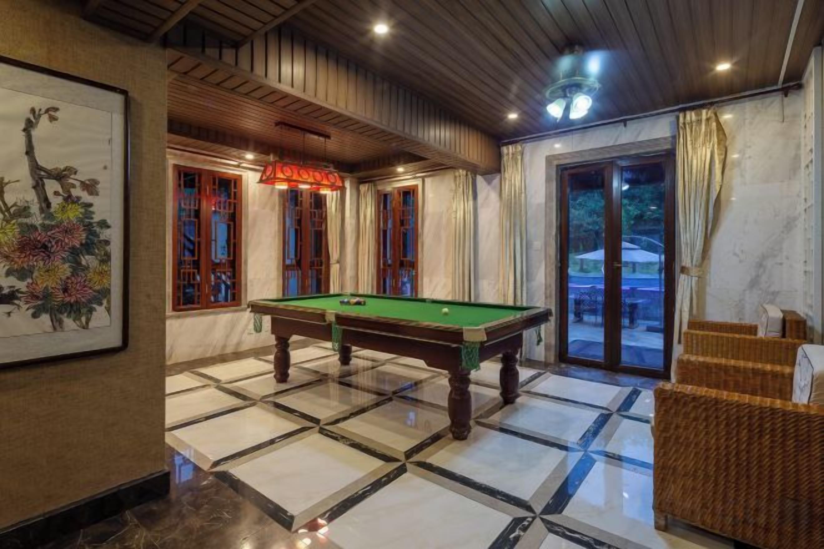 Una mesa de billar en una habitación con piso de baldosas, una obra de arte enmarcada en la pared y una silla de mimbre.