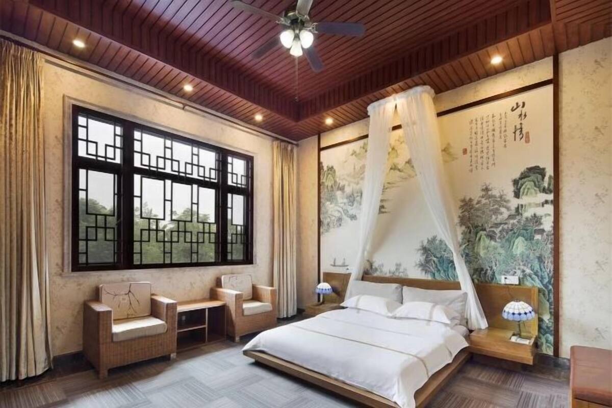 Une chambre à coucher comprenant un lit, deux chaises, une table de chevet, un ventilateur de plafond et une grande fenêtre ornée de motifs traditionnels chinois.