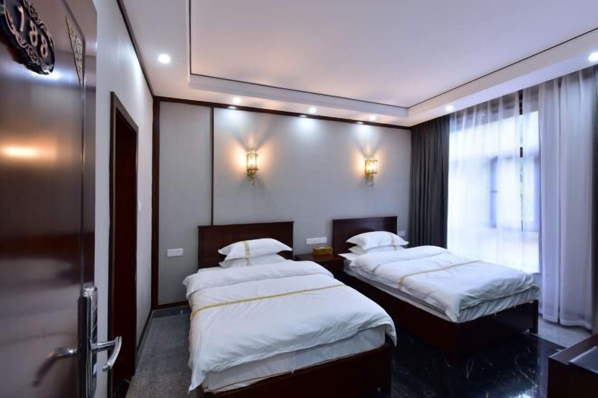 A hotel room with two beds, a large window with curtains, and wall-mounted lighting.