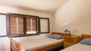 Une chambre de dortoir avec deux lits simples, une petite table de chevet en bois, un bureau et une fenêtre avec des stores.