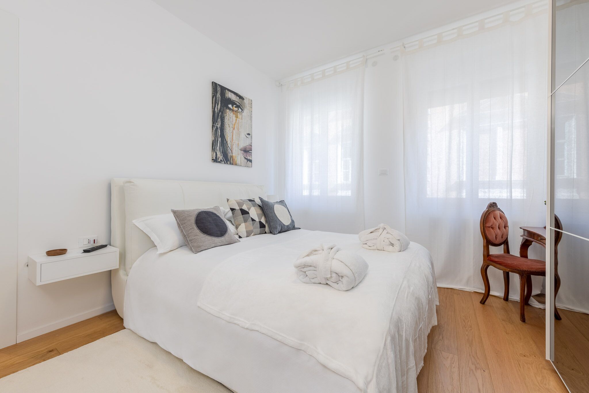 Une chambre à coucher avec un lit blanc, une table de chevet, une chaise et un tableau accroché au mur.