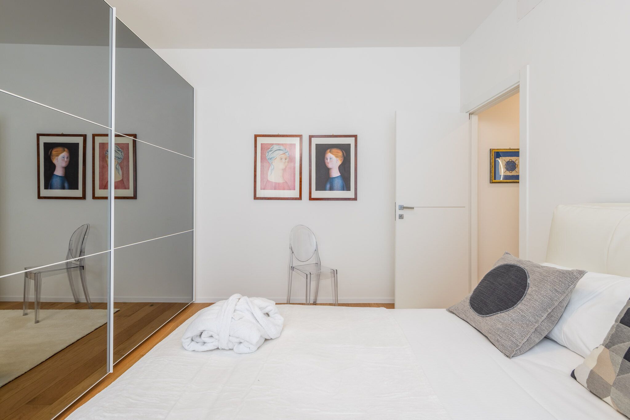 A modern bedroom with a large mirror, a bed with white bedding, a gray pillow, and two framed artworks on the wall.