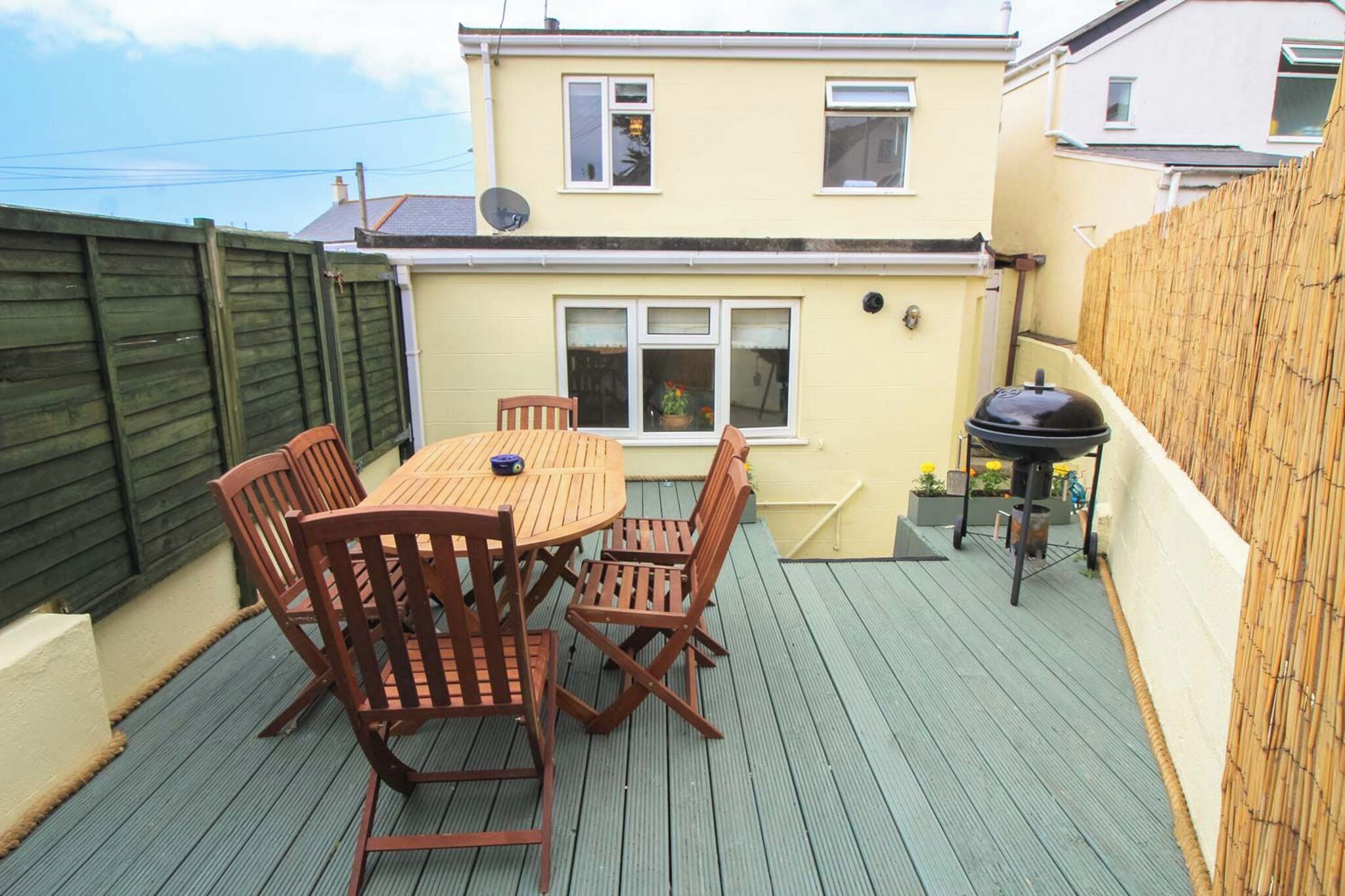 A patio with a wooden table and chairs, a barbecue grill, and a house in the background.