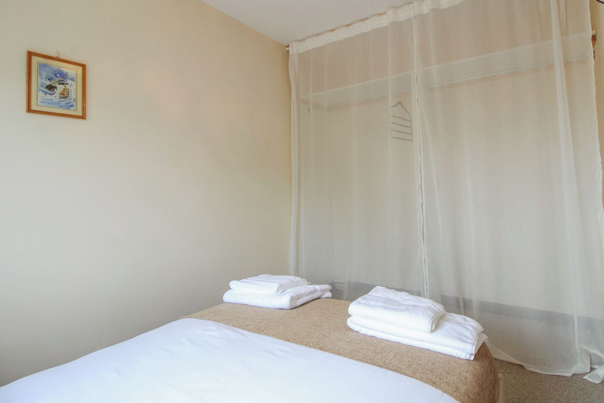 A hotel room with a bed, a small table with towels, a sheer curtain, and a framed picture on the wall.
