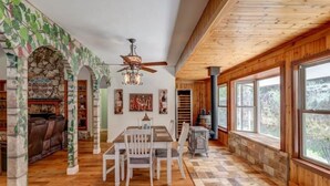 Una stanza spaziosa con soffitto in legno, ampie finestre, un camino e un'area pranzo con tavolo e sedie.