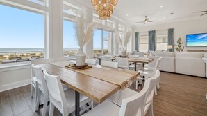 A spacious living room with a large wooden dining table, white chairs, and a white sofa. A large window offers a view of the beach. - Tides Beach House: Beachfront New Build- Sleeps 30, Pet Friendly, Priv. Pool (Cape San Blas)