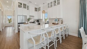 A modern kitchen with white cabinetry, a central island with bar stools, and a large refrigerator. - Tides Beach House: Beachfront New Build- Sleeps 30, Pet Friendly, Priv. Pool (Cape San Blas)