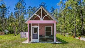 A small pink cabin with a pitched roof, a door, and a window, surrounded by a grassy area and trees. - Castaway Tiny Home in Bon Secour near Bon Secour River, 8 miles to sportsplex (Bon Secour)