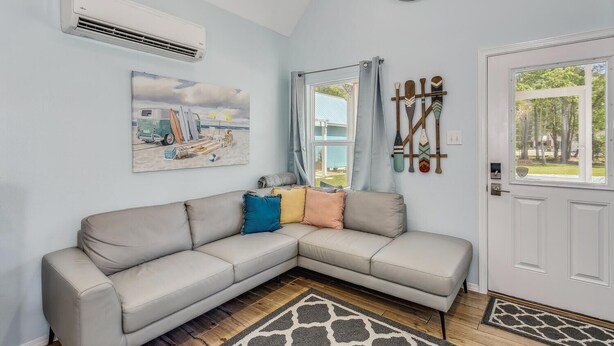 A living room with a beige sectional sofa, a painting of a beach scene, an air conditioning unit, a clock, and a door with a view of the outside. - Castaway Tiny Home in Bon Secour near Bon Secour River, 8 miles to sportsplex (Bon Secour)