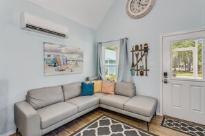 A living room with a beige sectional sofa, a painting of a beach scene, a clock, and a door with a view of the outside. - Castaway Tiny Home in Bon Secour near Bon Secour River, 8 miles to sportsplex (Bon Secour)