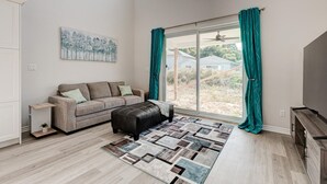 A spacious living room with a grey sofa, teal curtains, a black ottoman, and a large window offering a view of the outdoors. - Cozy, spacious family home w/ Arcade! 5 mins from Beach! (Port Stanley)