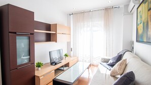 A modern living room with a built-in TV, a glass coffee table, a sofa with cushions, and a wooden cabinet with a built-in fridge. - Armilla Apartament by A3Rentals (Armilla)