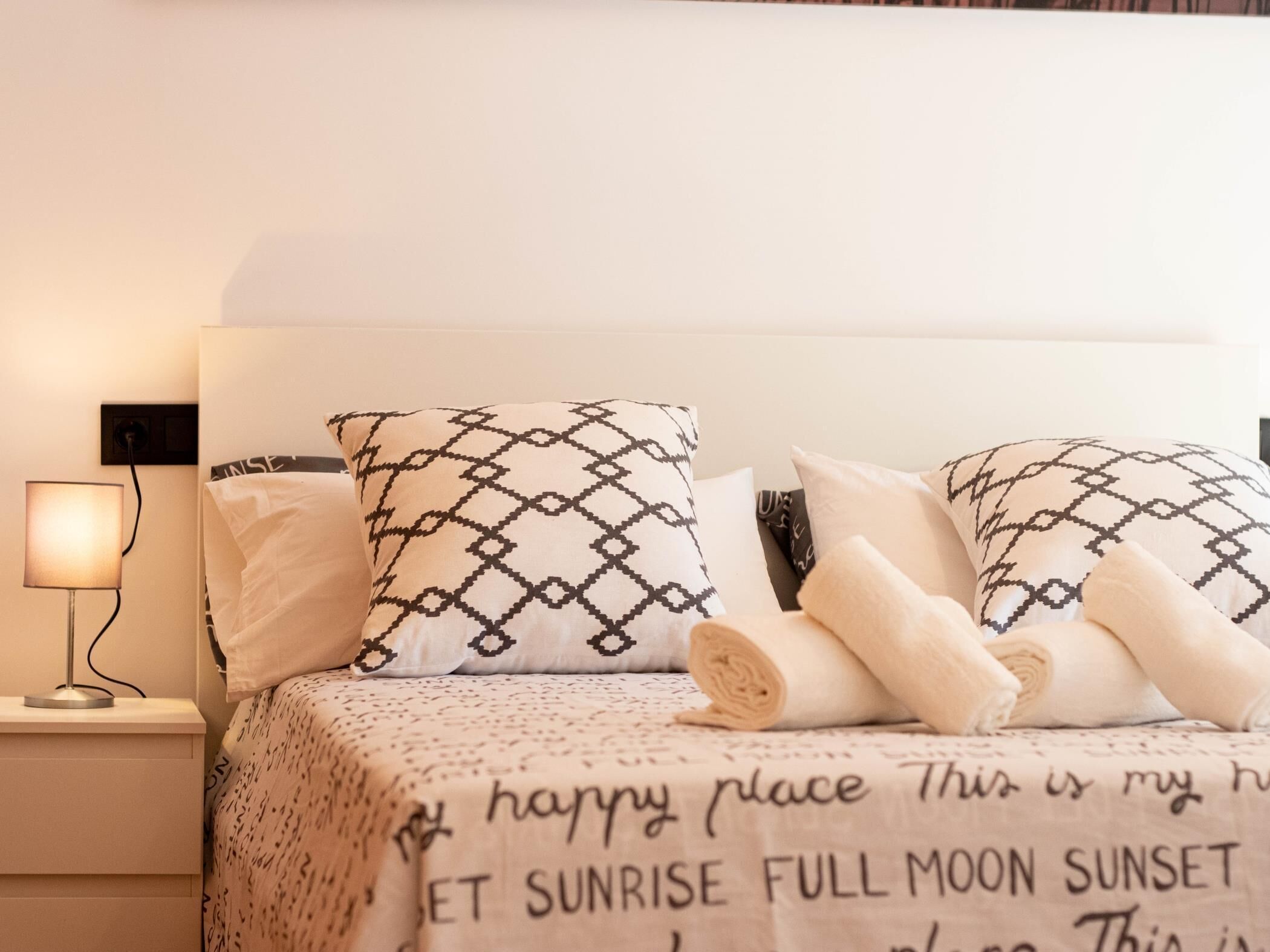 A neatly made bed with patterned pillows and rolled towels, a bedside lamp, and a wall-mounted power outlet.