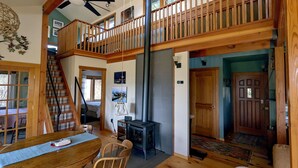 A spacious loft-style living area with a high ceiling, wooden staircase, and a dining area with a table and chairs.