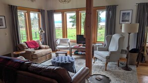 A cozy living room with a leather sofa, a tufted ottoman, and a striped armchair. A television is on a small table, and there are large windows with curtains.