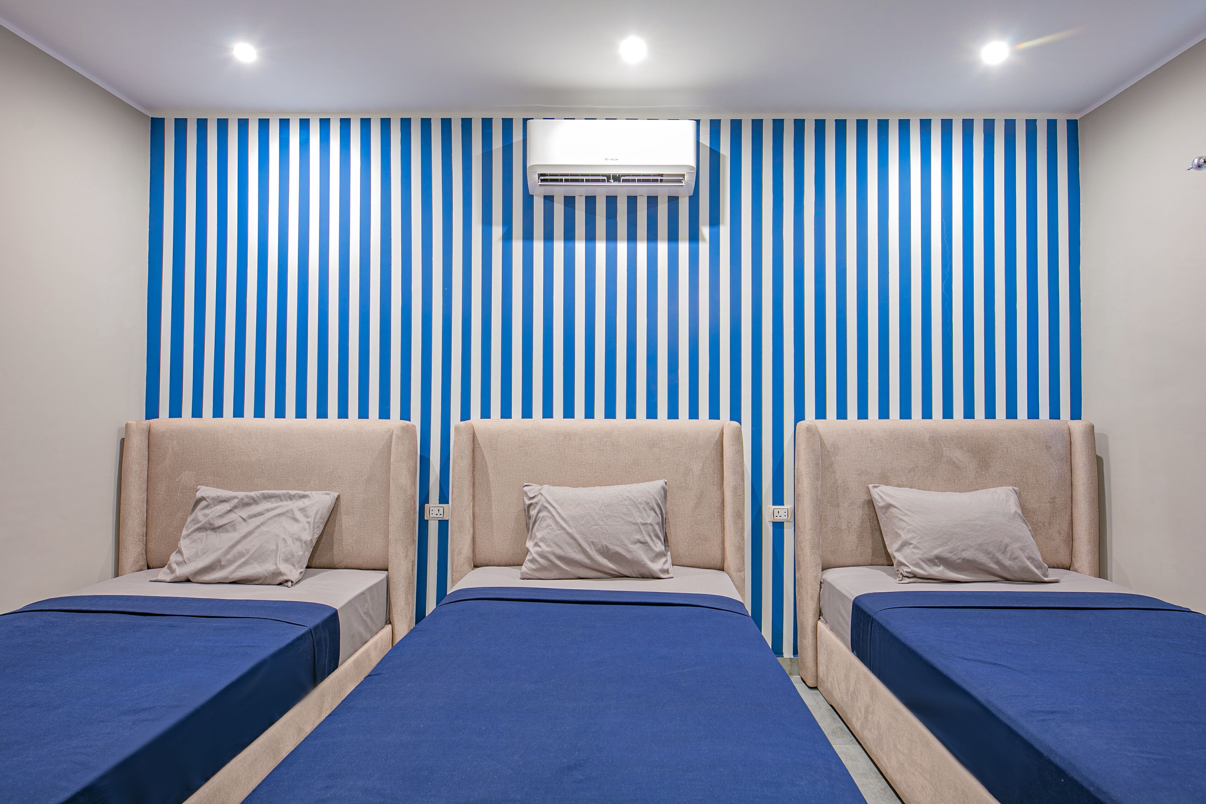 A room with three beds, each with a headboard and a pillow. There is a window with blinds and an air conditioning unit on the wall.