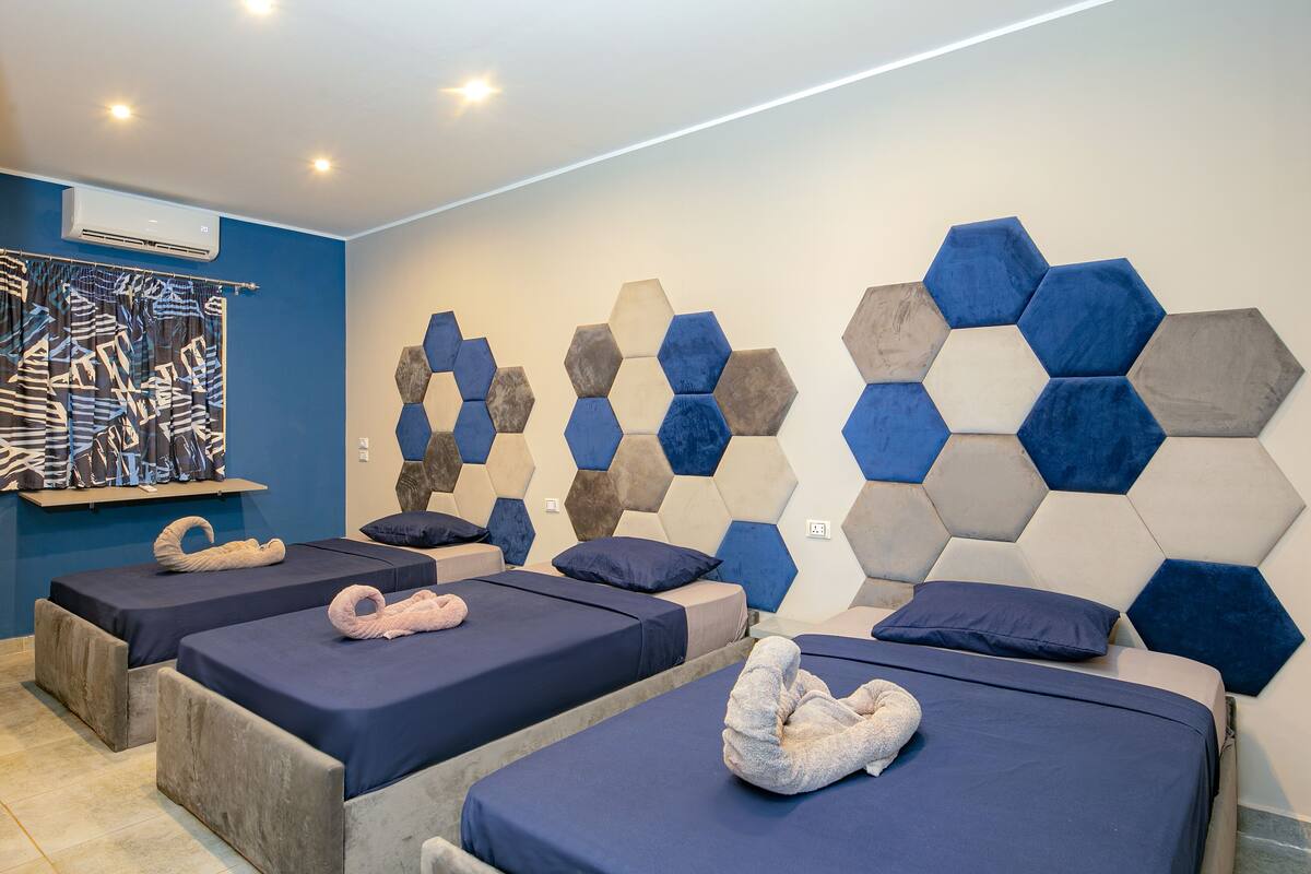 A hotel room with three beds, each with a dark blue bedspread and a towel. The walls are decorated with hexagonal patterns in various shades of blue and gray.