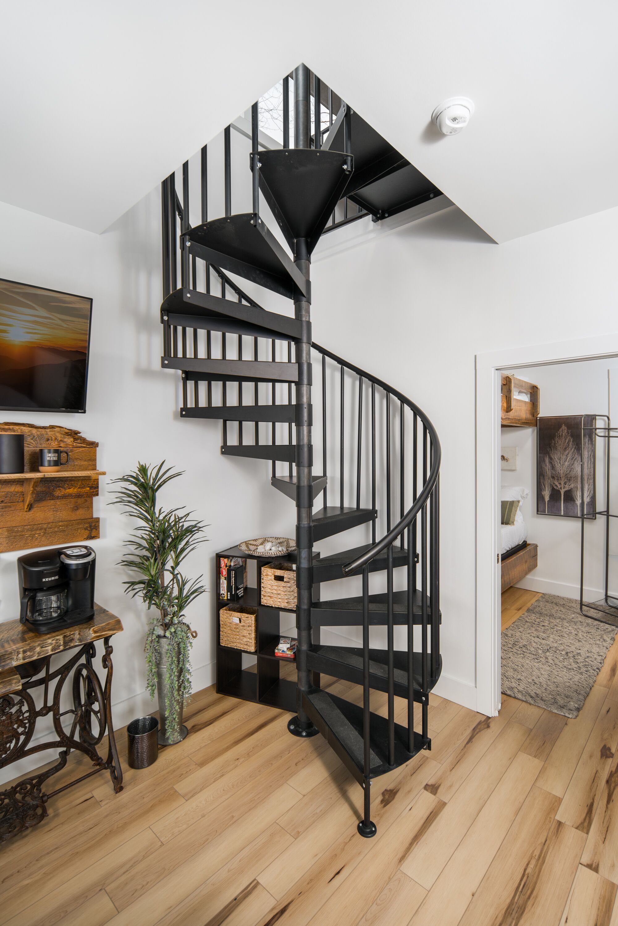 A modern spiral staircase with black railings in a room with wooden flooring, a coffee machine, a potted plant, and a painting on the wall.