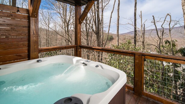 Una terraza de madera con un jacuzzi, rodeada por una cerca metálica y árboles.