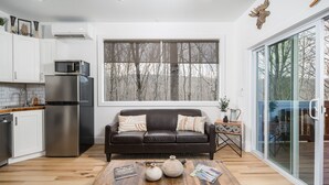 Una cocina moderna con armarios blancos, un refrigerador de acero inoxidable y un microondas. Hay un sofá de cuero marrón con cojines, una mesa de centro de madera con objetos decorativos y un ventanal con vista a árboles.