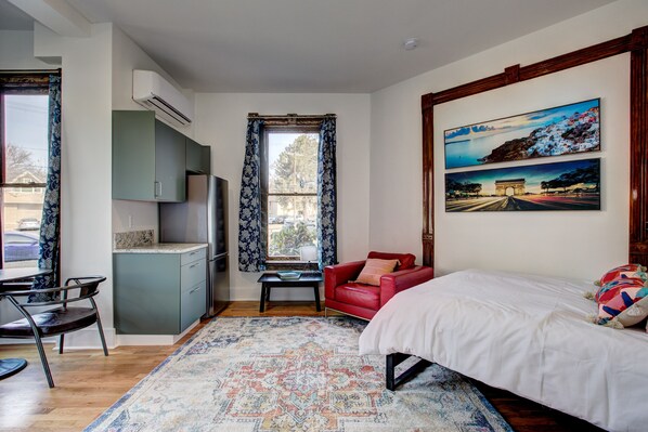 Un dormitorio con cama, sofá rojo, mesa de comedor con sillas, zona de cocina y un gran ventanal con cortinas.