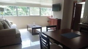 A modern living room with a sofa, a dining table with chairs, a small side table, a television mounted on the wall, and a view of greenery outside. - Nice apartment, Las Penas-Riverfront 1 (Guayaquil)