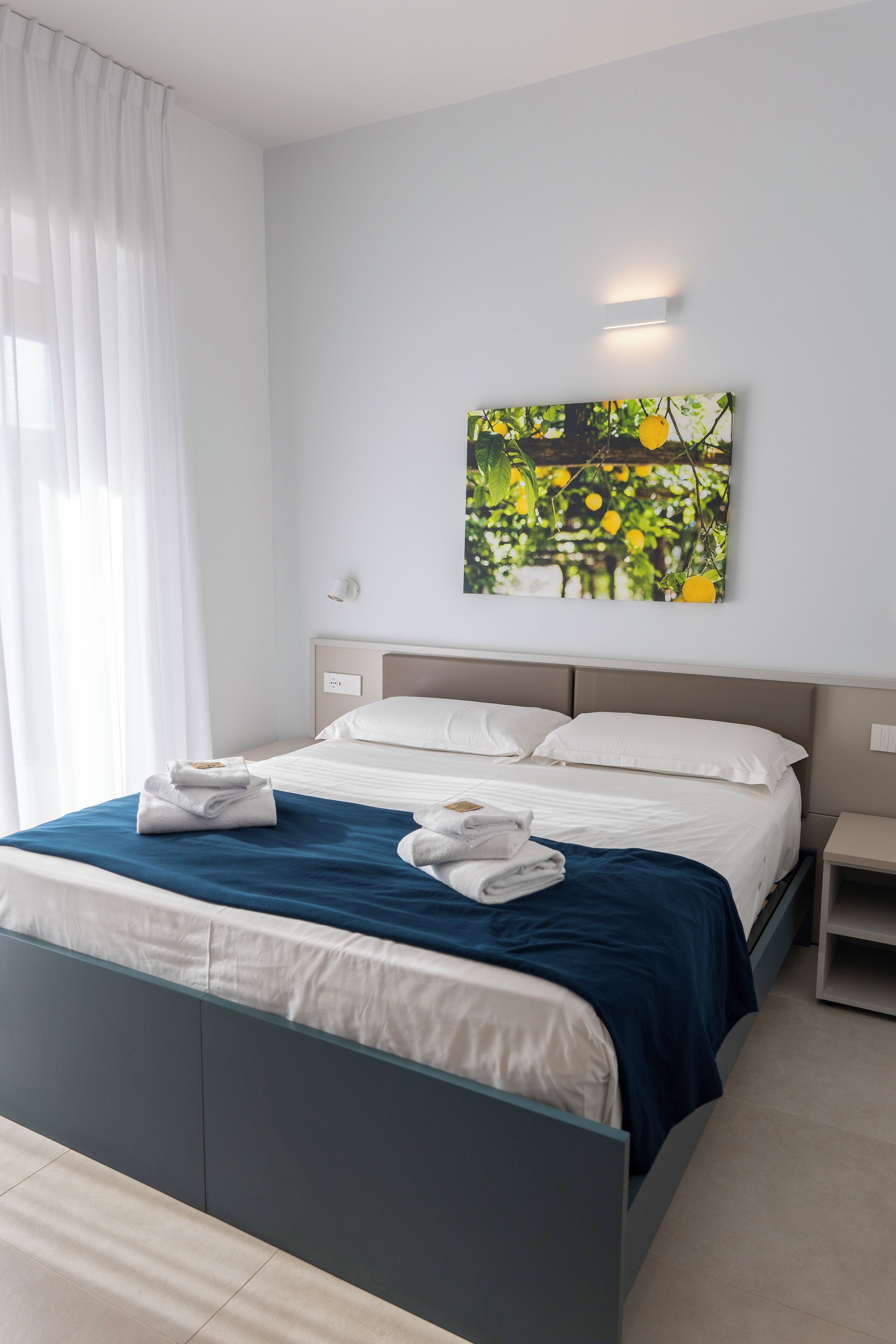 A modern hotel room with a bed, white curtains, a wall-mounted artwork, and a bedside table.