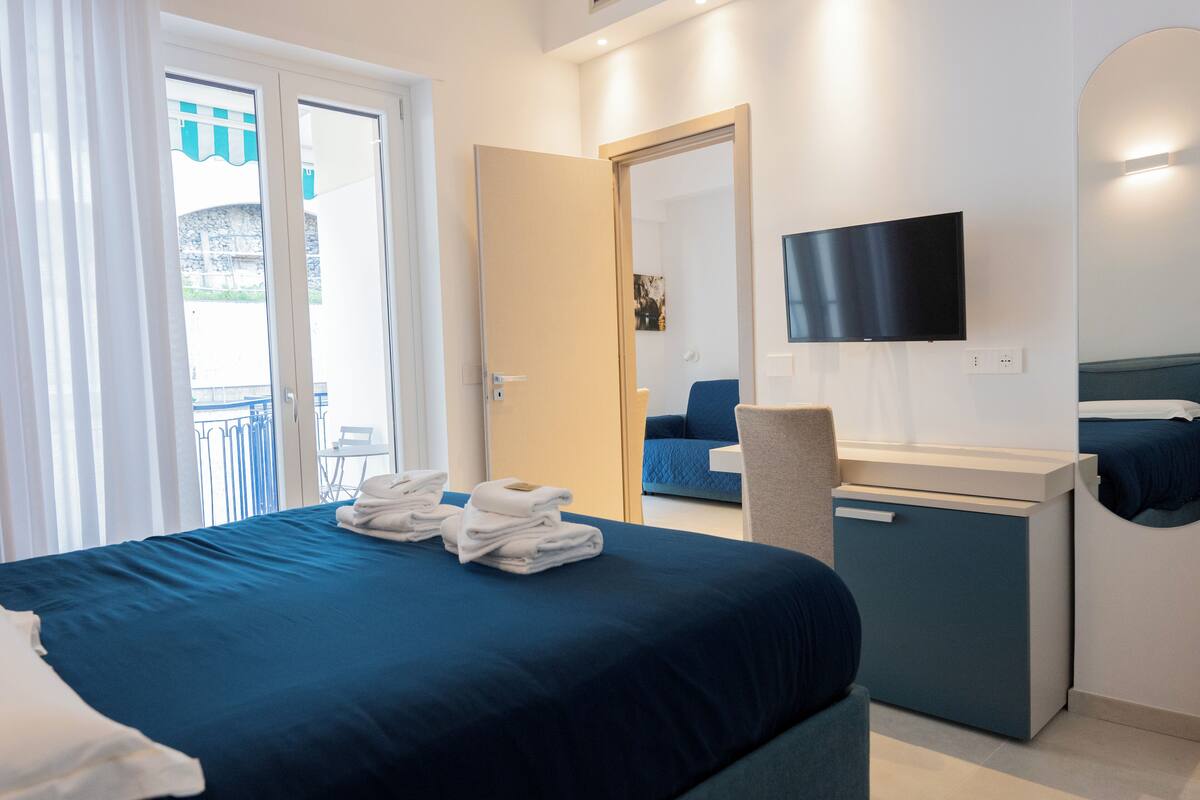 A hotel room with a bed, a desk, a chair, a TV, and a balcony with a view.