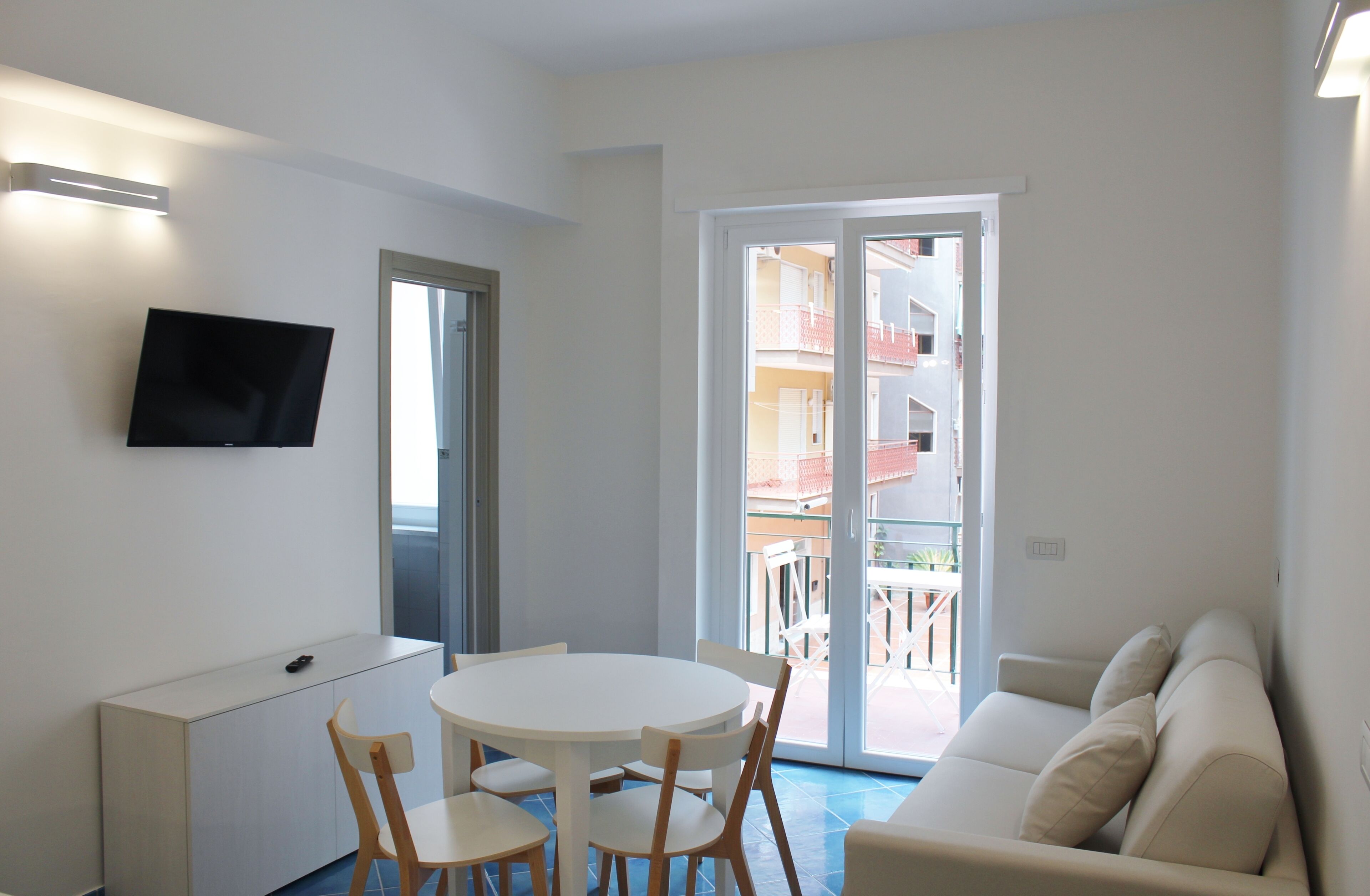 A modern living room with a flat-screen TV, a round dining table with chairs, a sofa, and a sliding glass door leading to an outdoor balcony.