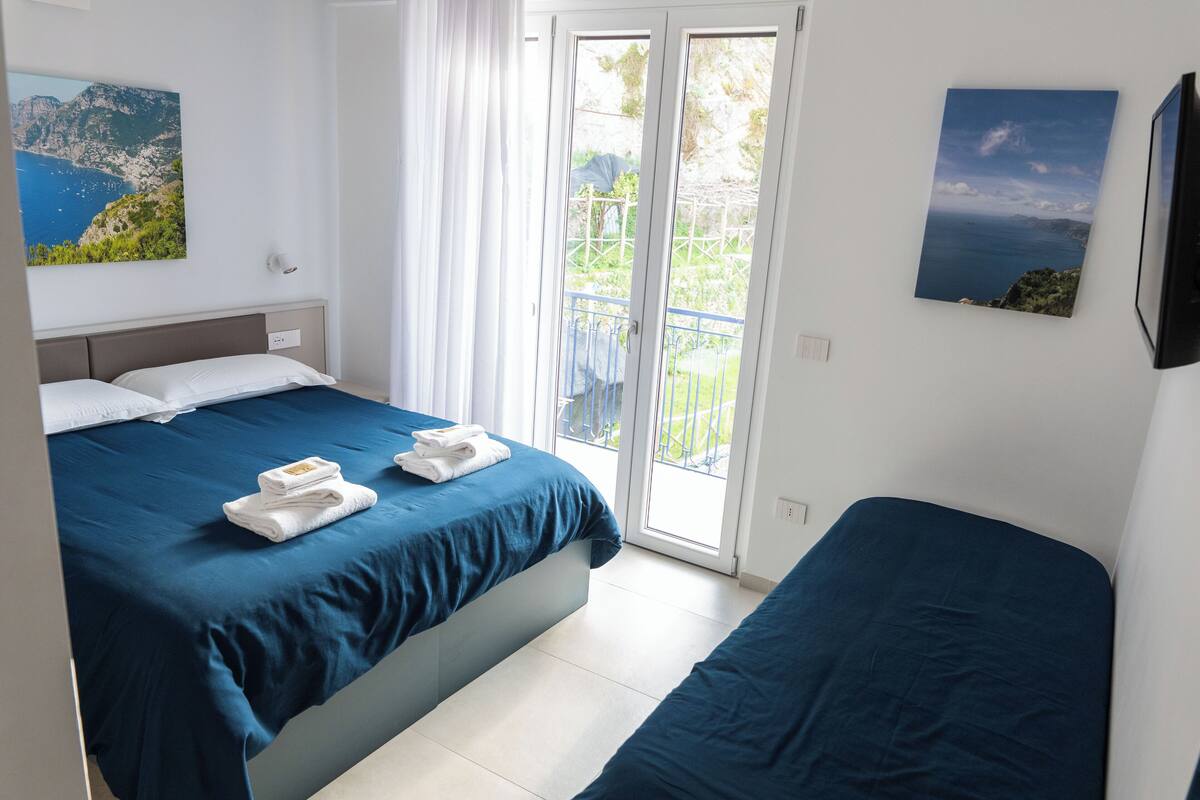 A bedroom with a bed, a sofa, a balcony with a view, and a painting on the wall.