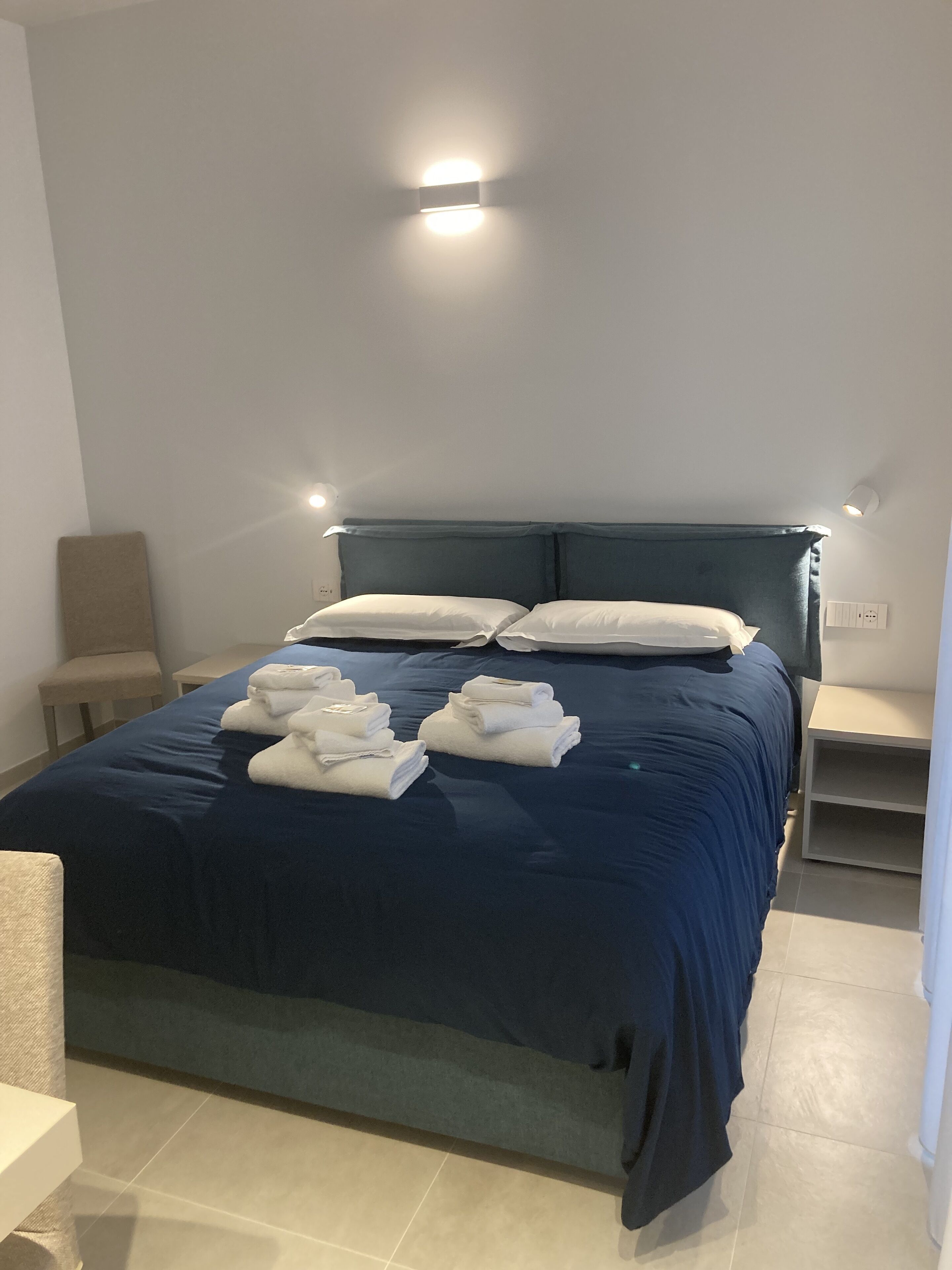 A neatly made bed with a dark blue bedspread and white pillows, a small bedside table, and a wall-mounted light fixture.