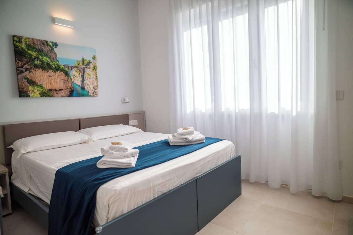 A hotel room with a bed, a painting of a coastal scene, and a window with sheer curtains.