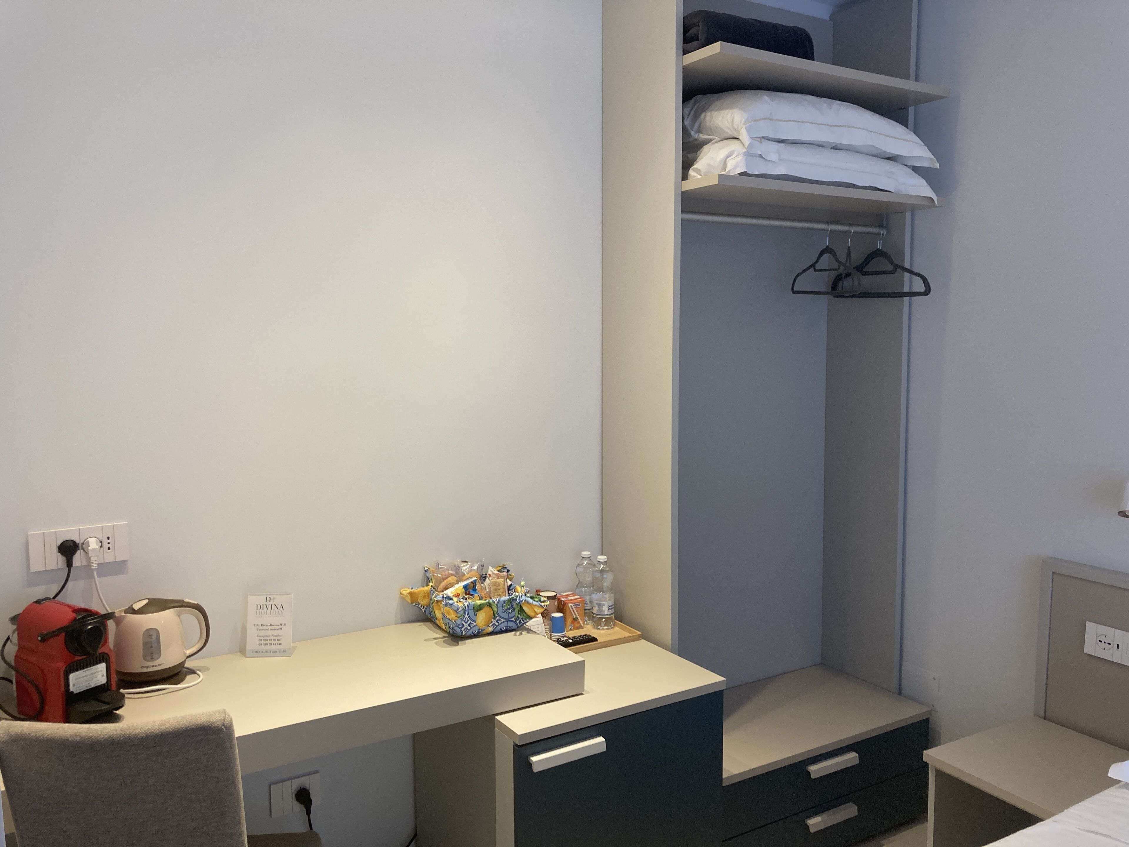 A compact hotel room with a desk, a chair, a bed, a wardrobe, and a shelf with towels.