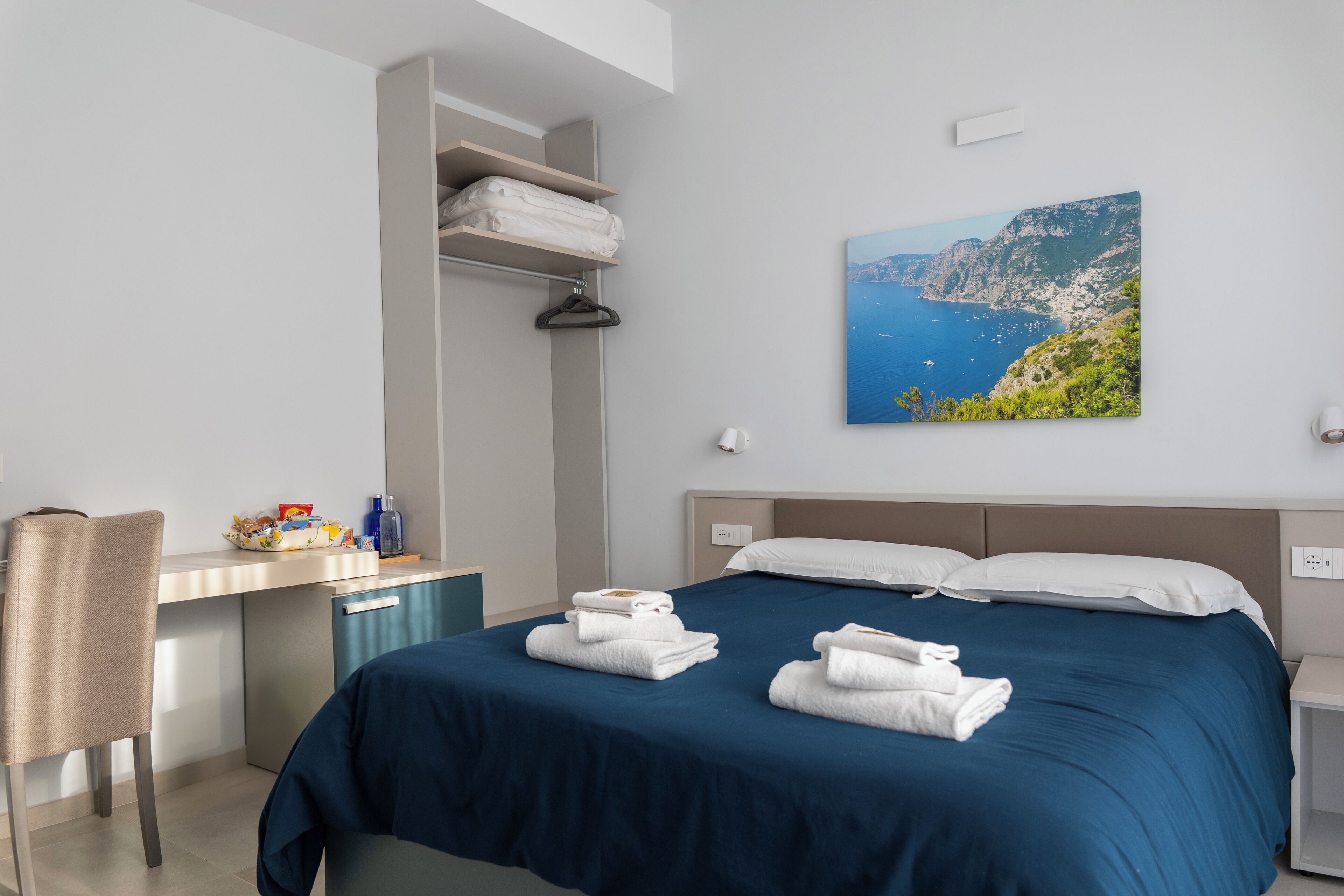 A hotel room with a bed, a desk, a chair, and a painting of a coastal landscape.