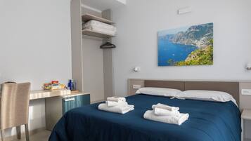 A hotel room with a bed, a desk, a chair, and a painting of a coastal landscape.