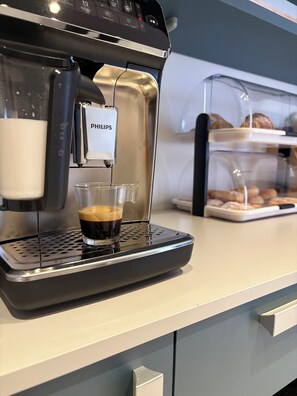 Eine Kaffeemaschine mit einem darunter stehenden Glas, einem Bildschirm und einer Auswahl an Gebäck im Hintergrund.