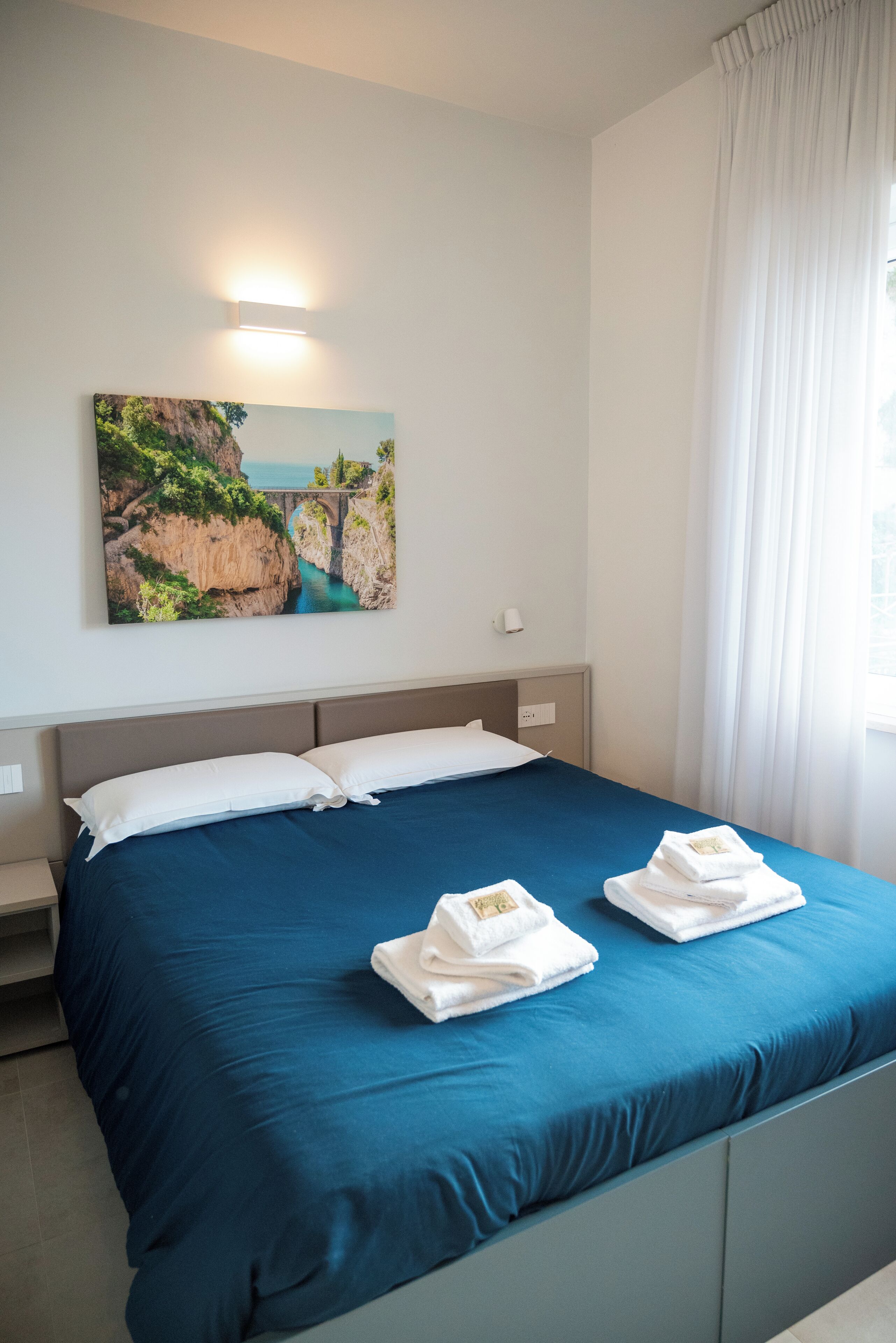 A hotel room with a large bed, a painting of a coastal landscape, and a window with curtains.
