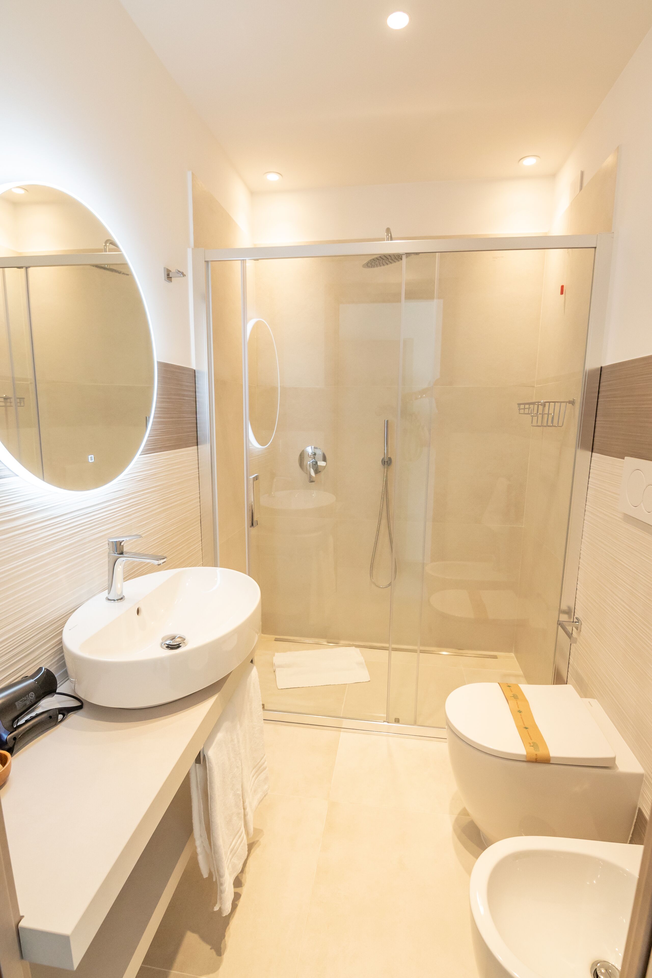 A modern bathroom with a glass-enclosed shower, a round mirror, a white sink, and a toilet with a towel rack.