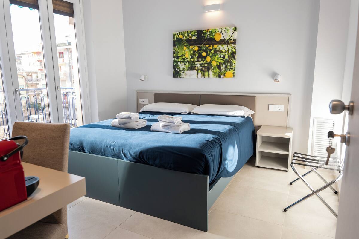 A modern hotel room with a bed, a desk, and a chair. There is a window with a view of buildings outside.