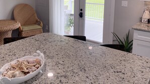A kitchen with a granite countertop, a wicker chair, and a view of the outdoors through a glass door. - The Yellowfin Inn- Down East Vacation Home with Sound Side Views (Gloucester)
