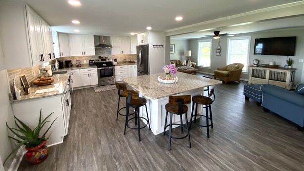 A spacious kitchen with white cabinets, a large island with bar stools, and a dining area with a blue sofa. - The Yellowfin Inn- Down East Vacation Home with Sound Side Views (Gloucester)