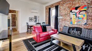 A modern living room with a red leather sofa, a black leather sofa, a wooden coffee table, and a black and white rug. There is a brick wall with a colorful abstract painting and a window with black curtains.