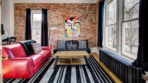 A room with a red leather sofa, a black and white striped rug, a wooden coffee table, and a large abstract painting on the brick wall.