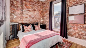 A bedroom with a brick wall, a large window with curtains, a bed with a red and white blanket, and a bedside table with a lamp.