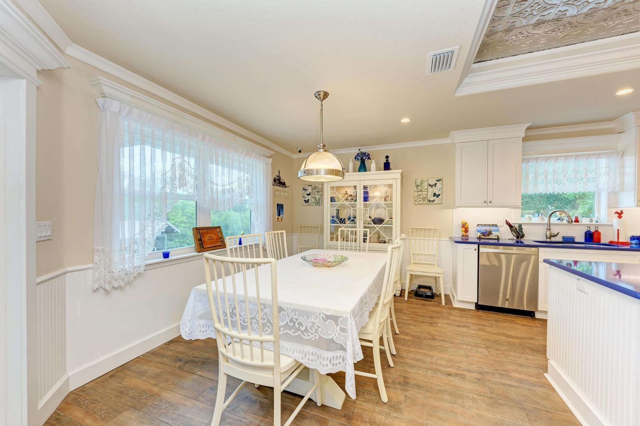 A dining area with a white table and chairs, a chandelier, and a view of the outdoors through large windows.