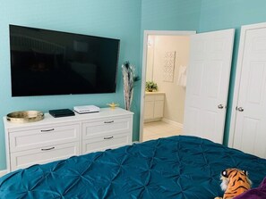 Un dormitorio con una televisión grande, un tocador blanco, una colcha azul y un peluche de tigre.