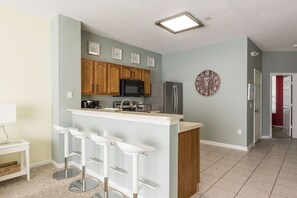 Cocina moderna con armarios de madera, refrigerador de acero inoxidable, microondas y una barra con taburetes blancos.