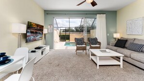 Ein Wohnzimmer mit Deckenventilator, Schwimmbadblick durch Glastüren, einem weißen Sofa und einem an der Wand montierten Fernseher.