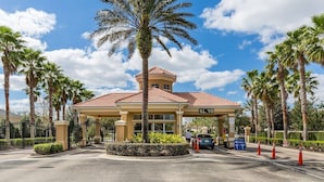 Entrada de hotel com palmeira, área de espera coberta e acesso por portão.