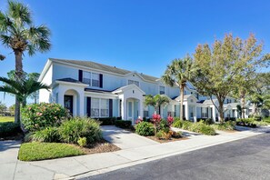 A row of white two-story houses with a uniform architectural style, featuring a central entrance with a covered porch, surrounded by well-maintained landscaping and palm trees.